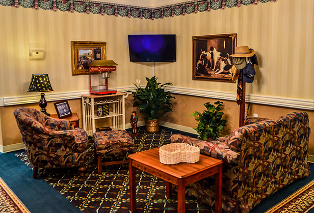 Common room seating area with patterned sofa and armchairs around a wooden table, a wall-mounted TV, plants, a popcorn machine and framed paintings.