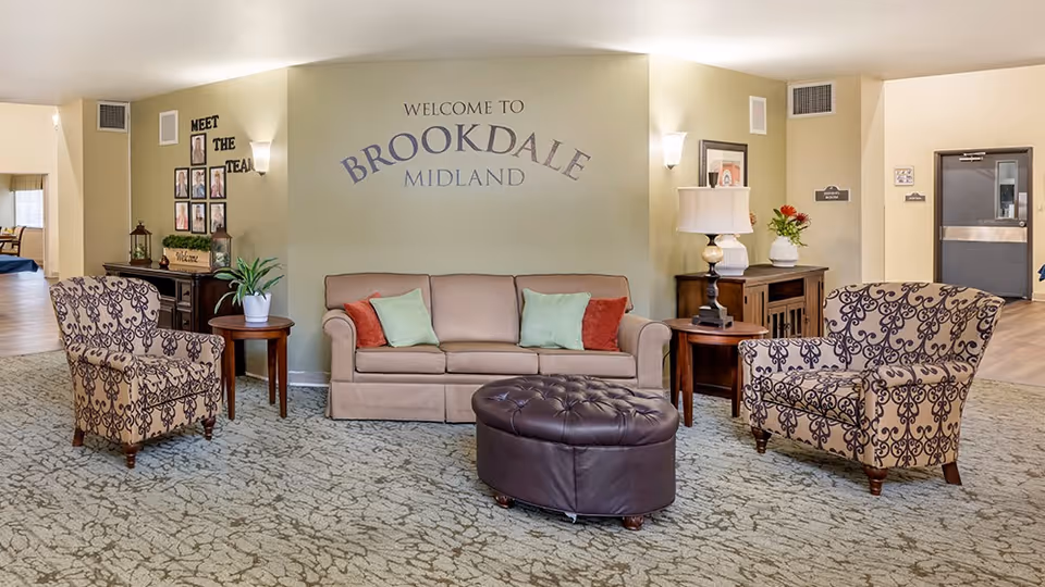 Lobby seating area with a sofa, patterned armchairs and ottoman under a 'Welcome to Brookdale Midland' wall sign.