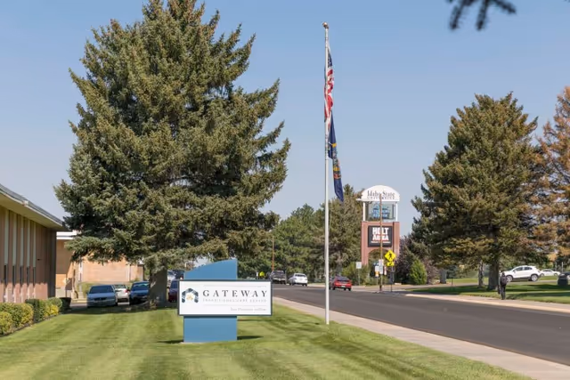 View of the Gateway Transitional Care Center sign on a grassy area next to a road, with large trees, parked cars, and a flagpole with two flags. In the background, there is a sign for Idaho State University and a clear blue sky.