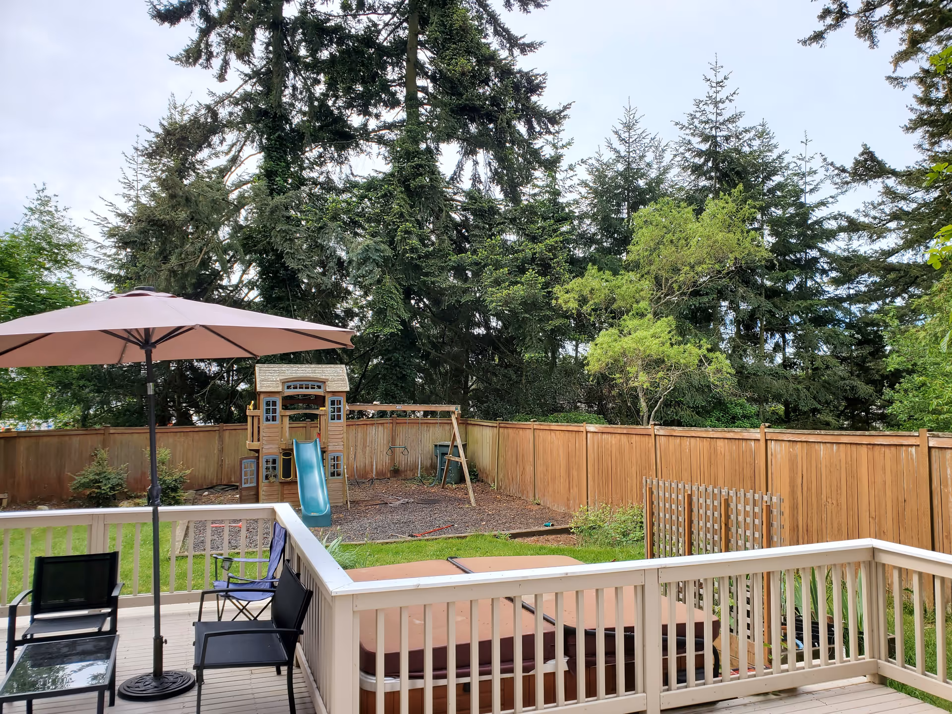 View of a fenced backyard with a wooden playset featuring a slide and swings, surrounded by tall evergreen trees. In the foreground, there is a wooden deck with black chairs, a glass table, a large beige patio umbrella, and a covered hot tub.