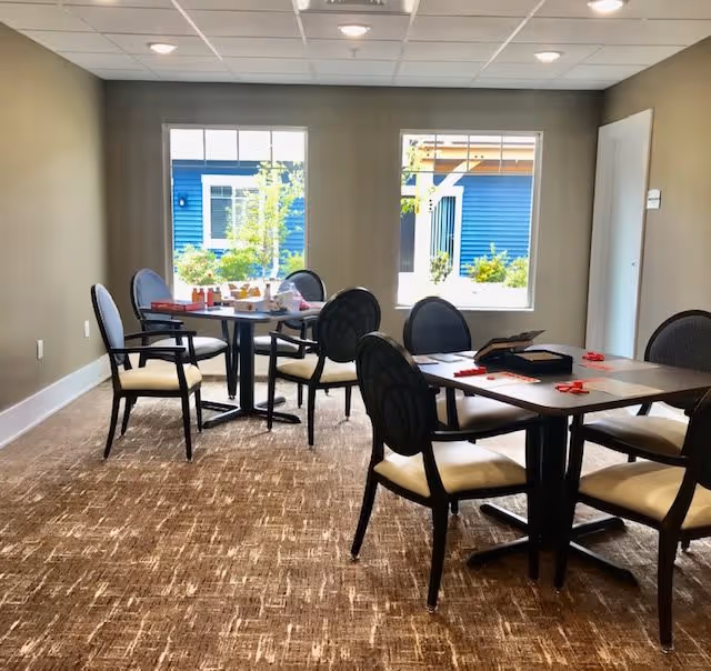 Interior room with two tables and several chairs arranged around them. The tables have some items on them, including a game board and game pieces. Two large windows show an outdoor view with a blue building and greenery.