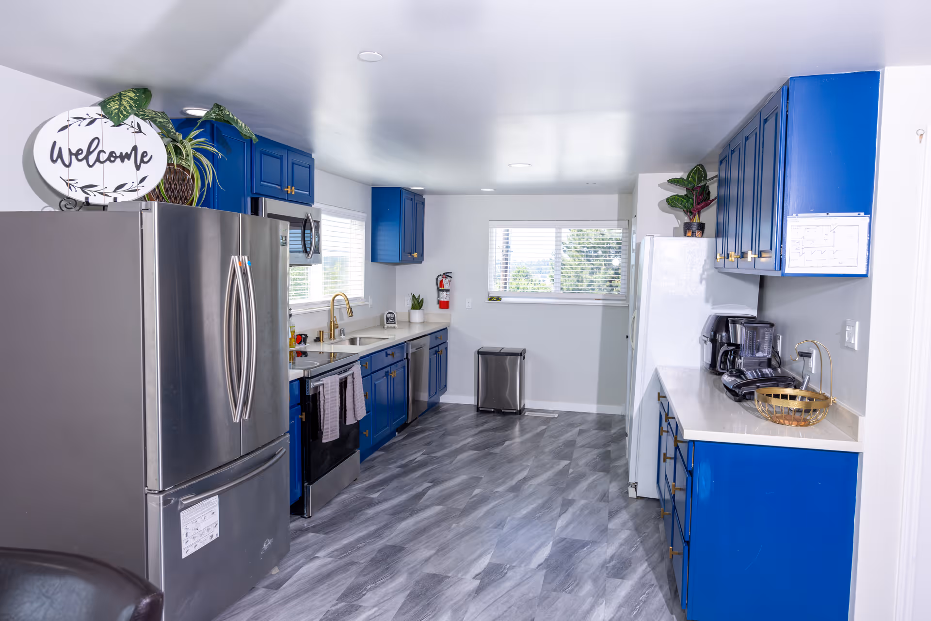 A modern kitchen with blue cabinets and stainless steel appliances including a refrigerator, stove, and dishwasher. The kitchen has a white countertop, a double sink with a gold faucet, and a window with blinds letting in natural light. There are plants on top of the refrigerator and cabinets, a coffee maker on the counter, and a 'Welcome' sign on top of the refrigerator. The floor has a gray patterned design.