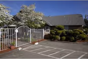 Single-story brown building entrance with a white metal gate, trimmed shrubs, and marked parking spaces under a clear blue sky.