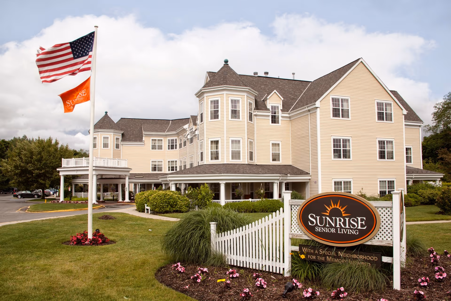 Exterior front view of the Sunrise Senior Living building with flagpoles, landscaped lawn, and the facility sign in the foreground.
