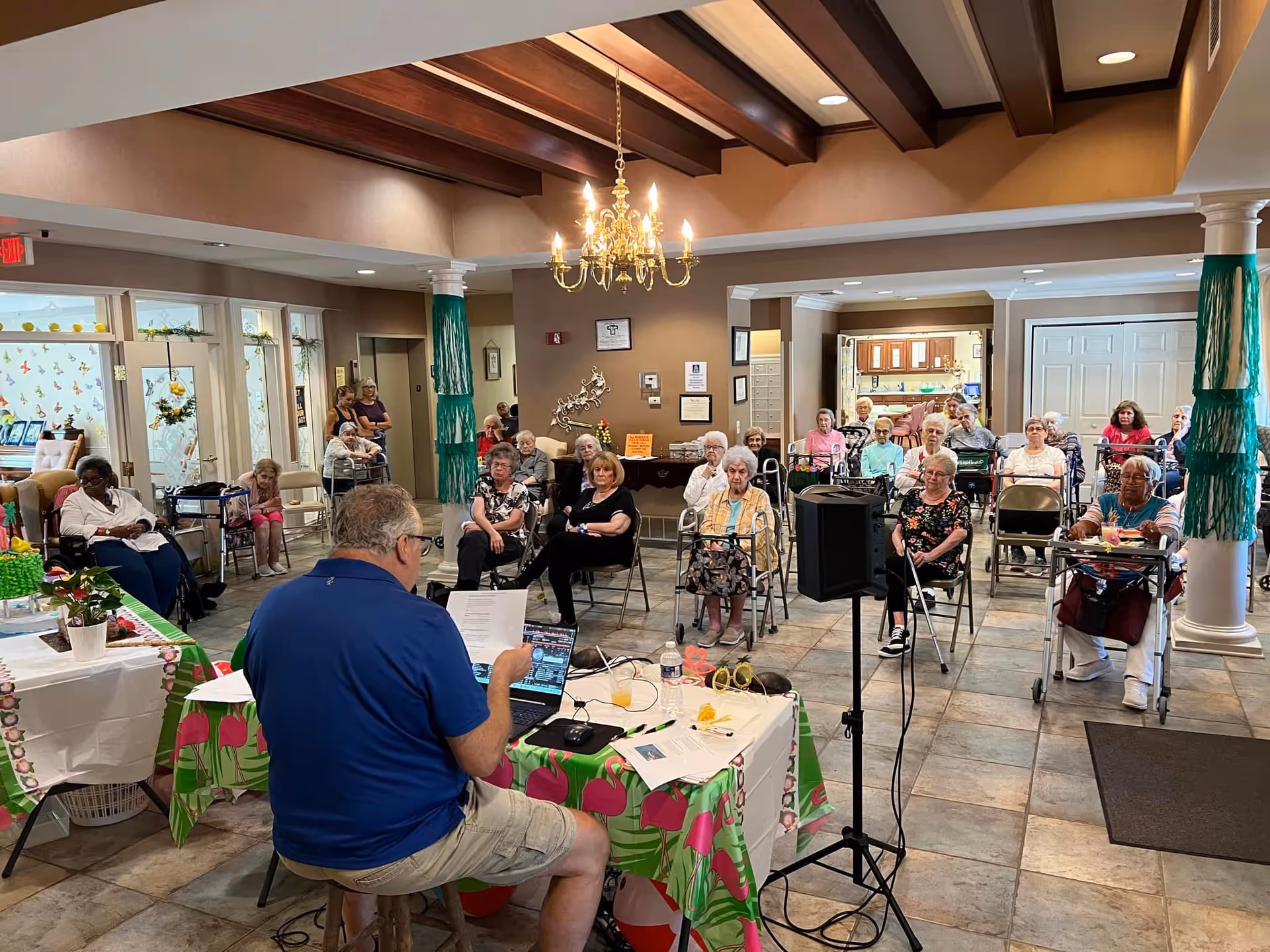 A group of elderly residents seated in chairs and using walkers in a spacious common area with tiled floors and wooden ceiling beams. A man in a blue shirt and shorts is seated at a table with a laptop and papers, facing the residents and appearing to lead an activity or presentation. The room is decorated with green and pink tablecloths and has a chandelier hanging from the ceiling.