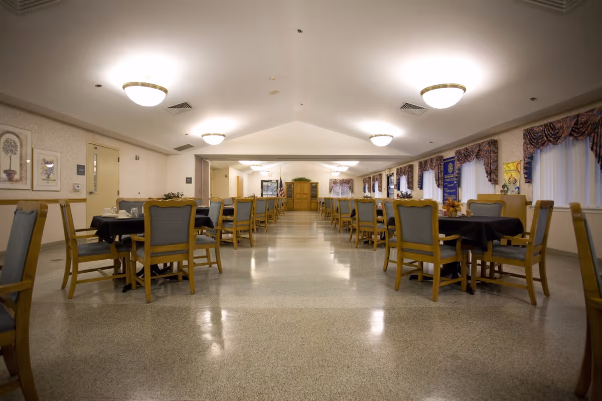 Spacious dining room with rows of tables and chairs under ceiling lights in a senior living facility.