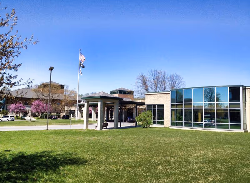Front exterior of a healthcare facility with a covered entrance, flagpoles, large glass windows, and a grassy lawn under a clear blue sky.