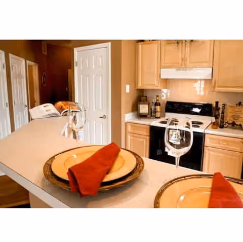 View of a kitchen area with light wood cabinets, a white stove, and a countertop with place settings including plates, red napkins, and wine glasses.