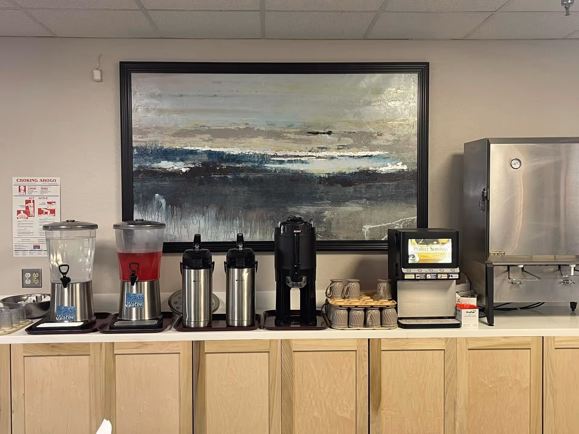 A beverage station with dispensers containing water and a red drink, two stainless steel airpots, a black coffee dispenser, a stack of beige mugs, a small machine labeled Perfect Serving, and a large stainless steel appliance on a countertop with light wood cabinets below. A large abstract painting with blue, gray, and beige tones hangs on the wall above the station. A choking emergency instruction poster is visible on the left wall.
