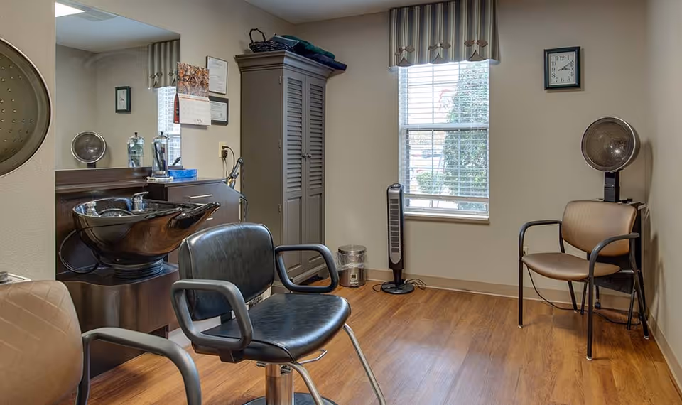 Interior of a small hair salon area with a black salon chair in front of a black wash basin, a large mirror on the wall, a gray cabinet, a window with blinds and a striped valance, a standing fan, a clock on the wall, and two additional chairs for waiting.