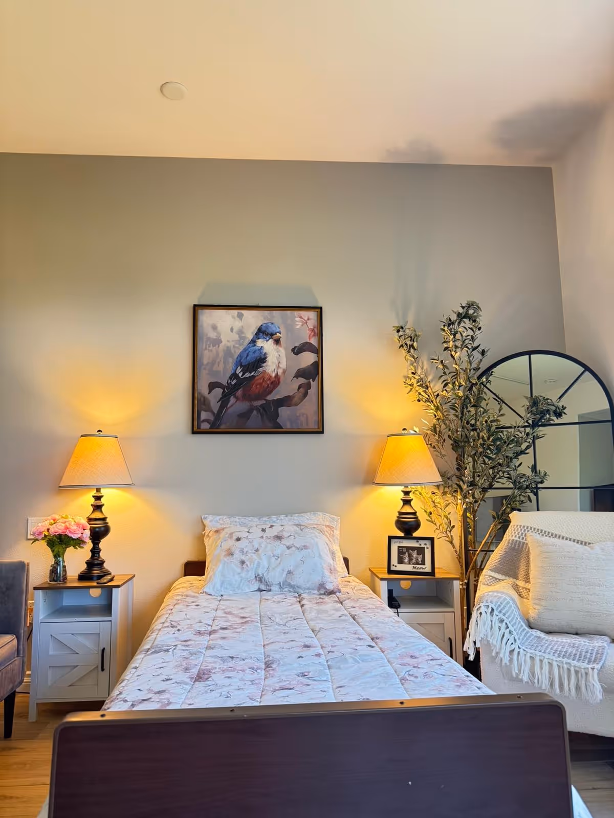 A cozy bedroom with a single bed covered in a floral quilt and matching pillow. On either side of the bed are white nightstands with black lamps emitting warm light. Above the bed hangs a framed painting of a bird. To the right, there is a large leafy plant and a round mirror behind a white armchair draped with a textured throw blanket and a pillow. A small vase with pink flowers is on the left nightstand.