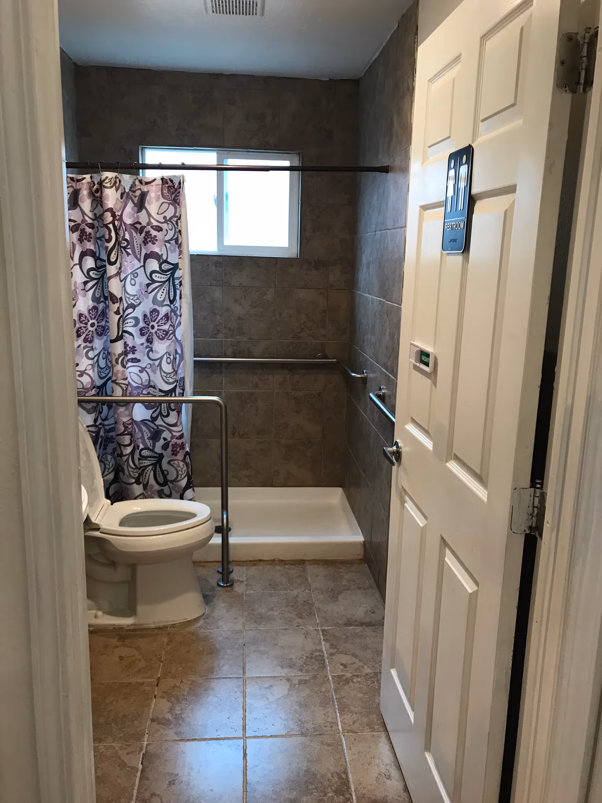 A small bathroom with a toilet and a walk-in shower with a floral patterned shower curtain. The walls and floor are tiled in a brownish color. There are metal grab bars installed near the toilet and inside the shower for accessibility. A small window is above the shower area, and the door has a sign indicating it is a restroom.