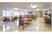 A spacious and well-lit common area with multiple tables and chairs arranged for seating. Large windows allow natural light to fill the room, and ceiling lights provide additional illumination. The floor has a light-colored pattern, and there are plants and decorative elements along the walls.