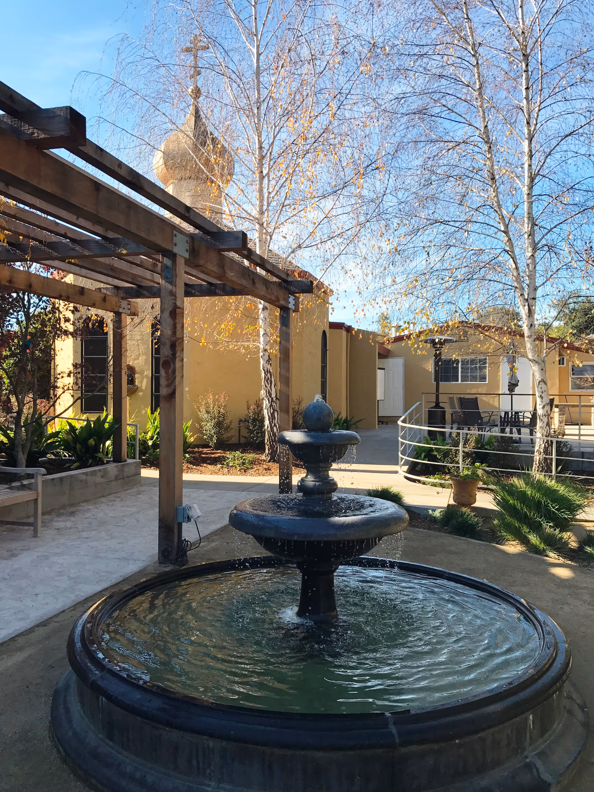 Outdoor courtyard area with a three-tiered water fountain in the foreground, surrounded by trees with sparse leaves and a wooden pergola on the left. In the background, there is a beige building with a dome topped by a cross, and a patio area with chairs and a table.