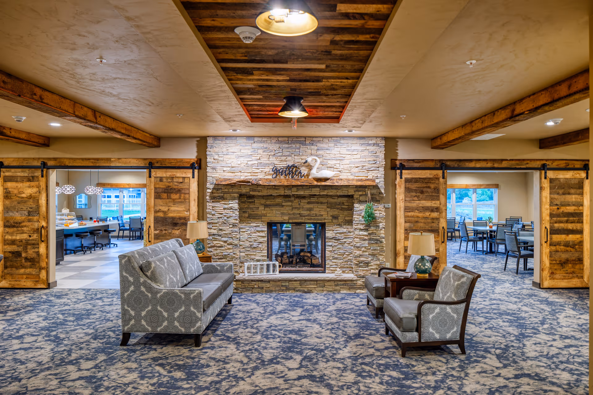 A cozy senior living common area with a stone fireplace in the center, flanked by two wooden barn-style sliding doors. The room features patterned carpet, a sofa, two armchairs, and side tables with lamps. Through the open doors, dining areas with tables and chairs are visible.