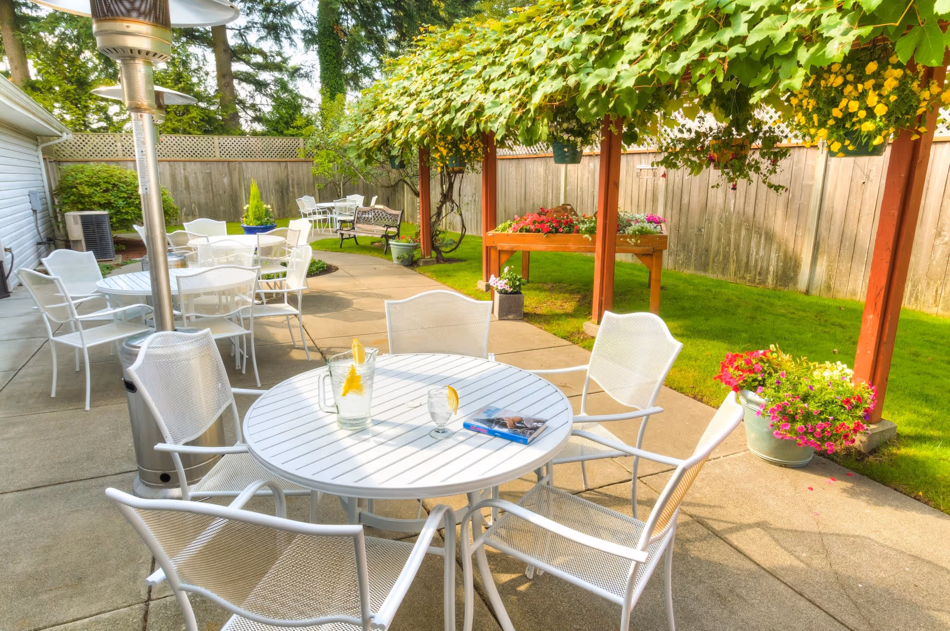 Outdoor patio area with white metal tables and chairs arranged on a concrete surface. A pitcher of water with lemon slices and a glass are on the nearest table, along with a magazine. There are hanging flower baskets and potted plants around the patio, with a wooden fence and green grass in the background. Trees and shrubs provide additional greenery.