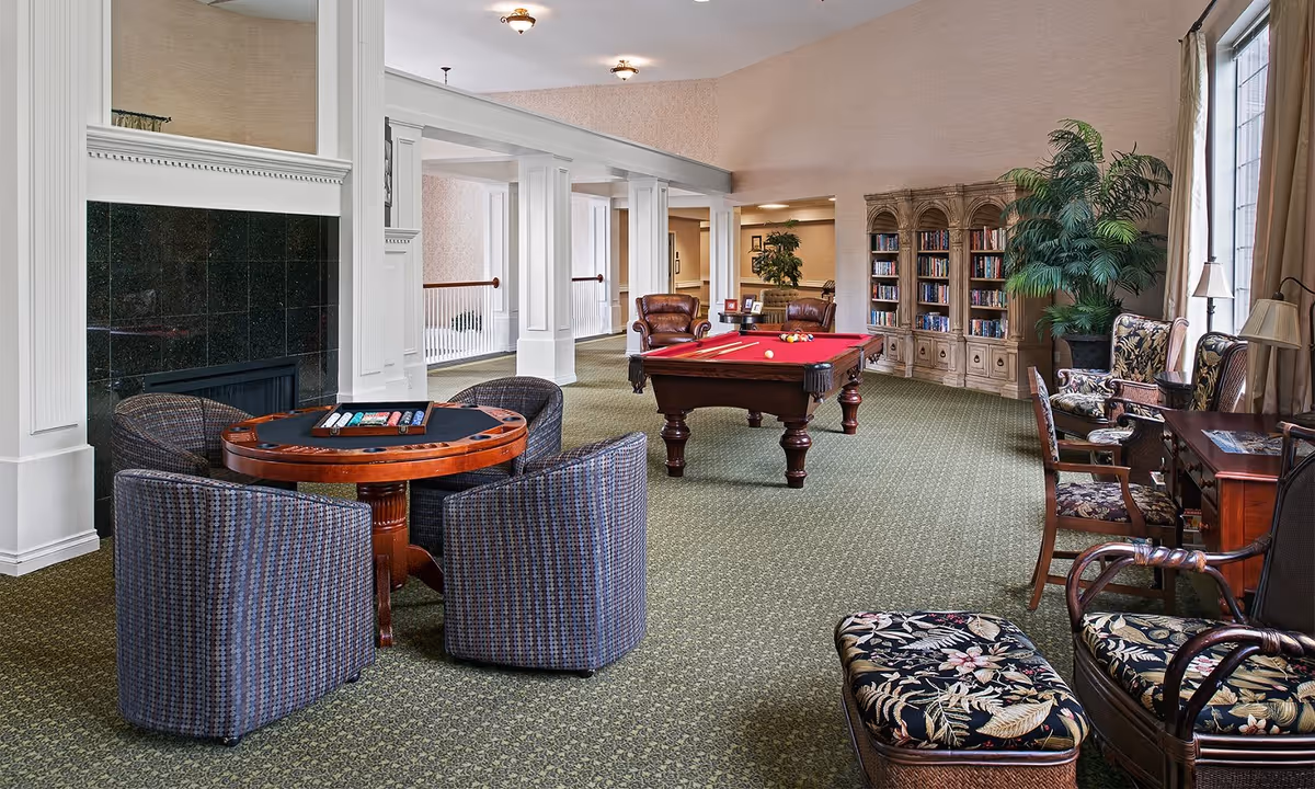 A spacious and well-lit common area featuring a pool table with red felt, a round card table with four chairs, a fireplace with a black tile surround, a large bookcase filled with books, several armchairs, and large windows with curtains allowing natural light to enter.