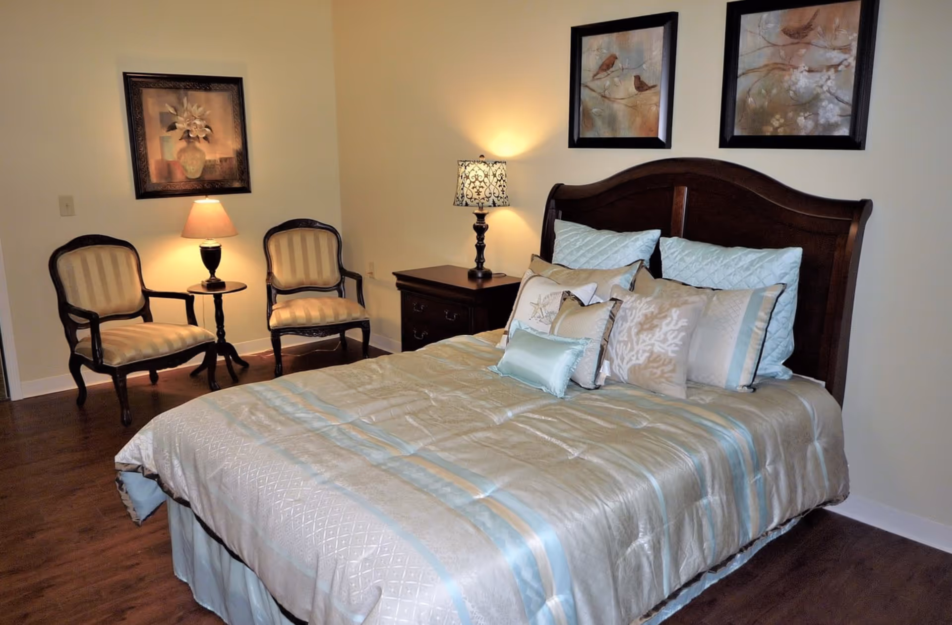 A bedroom with a large bed covered in a light blue and beige comforter and multiple decorative pillows. The bed has a dark wooden headboard. To the right of the bed is a dark wooden nightstand with a decorative lamp. On the left side of the room, there are two upholstered armchairs with a small round table and a lamp between them. The walls are light-colored and decorated with framed artwork.