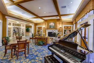 A bright senior living lounge with a grand piano, seating, and decorative fixtures.