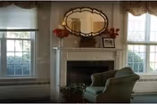 A cozy interior room featuring a green upholstered armchair positioned near a white fireplace with decorative molding. Above the fireplace is an ornate oval mirror, flanked by red floral arrangements and a framed photograph. Two large windows with white blinds and valances allow natural light to fill the room.