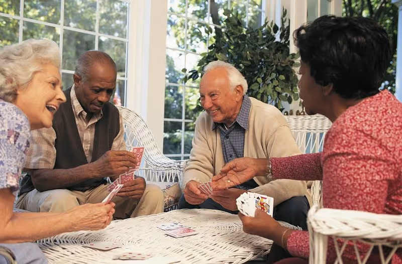 Four elderly people sitting around a white wicker table playing cards in a bright room with large windows and green plants in the background. They appear to be enjoying each other's company and smiling.