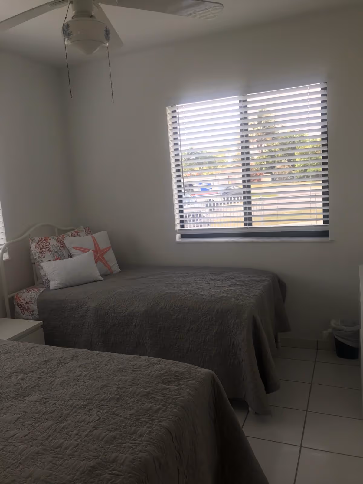 A small bedroom with two twin beds covered in gray quilts. One bed is positioned against the wall under a window with horizontal blinds. The bed near the window has decorative pillows with coral and starfish designs. The room has white tiled floors, white walls, a ceiling fan with a light, and a small trash bin in the corner.