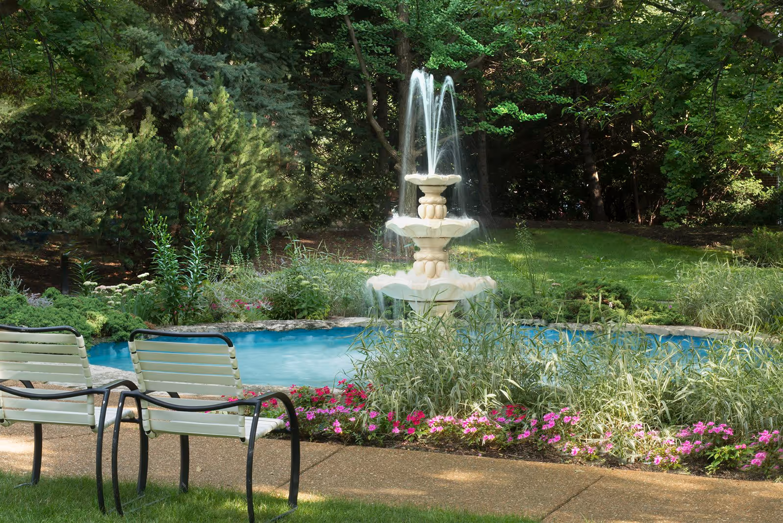 A peaceful outdoor garden area with a decorative three-tiered water fountain surrounded by greenery and pink flowers. Two white metal chairs with black armrests are positioned on a paved pathway facing the fountain. Tall trees and lush plants create a serene natural backdrop.