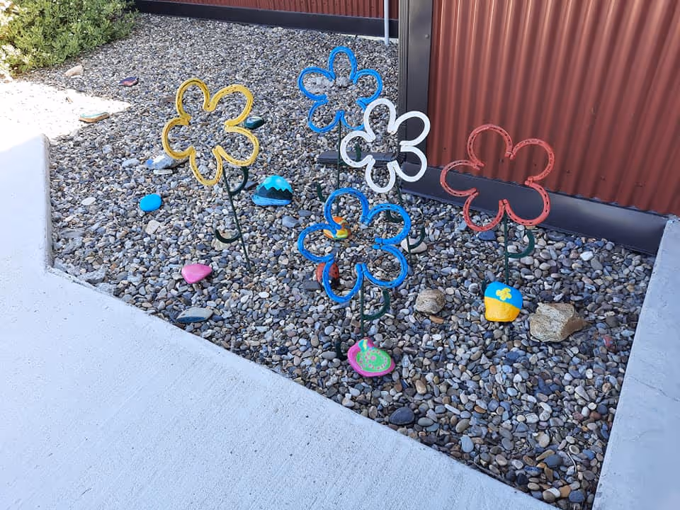 A small outdoor garden area with decorative metal flowers in yellow, blue, white, and red colors planted in a bed of small rocks. The garden is bordered by a concrete walkway and a red corrugated metal wall.