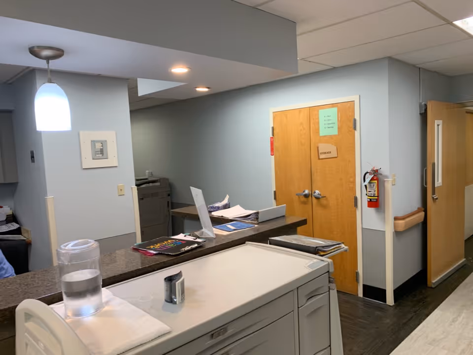 Interior view of a reception or nurse station area in a senior living facility. The image shows a countertop with a water pitcher, a cup, some papers, and office supplies. Behind the counter, there is a printer and a double wooden door labeled 'Storage'. The walls are painted light blue, and there is a fire extinguisher mounted on the wall next to the door. The floor has a dark wood pattern, and there is a hallway leading to other rooms.