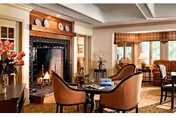 A cozy living room with a lit fireplace surrounded by a wooden mantel displaying decorative plates. Two upholstered chairs and a small round table are positioned in front of the fireplace. The room features large windows with plaid valances, a sofa, and additional seating, creating a warm and inviting atmosphere.