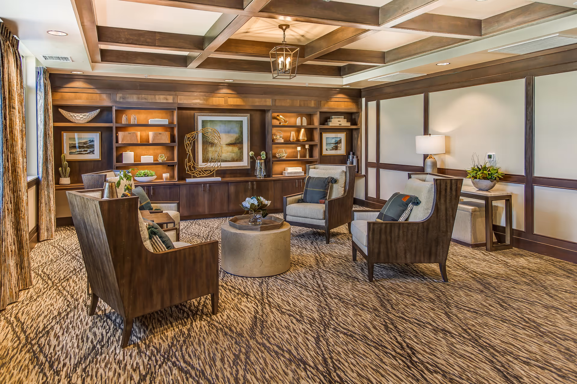 Cozy senior living lounge with armchairs arranged around a round coffee table, built-in wooden shelving and a coffered ceiling.