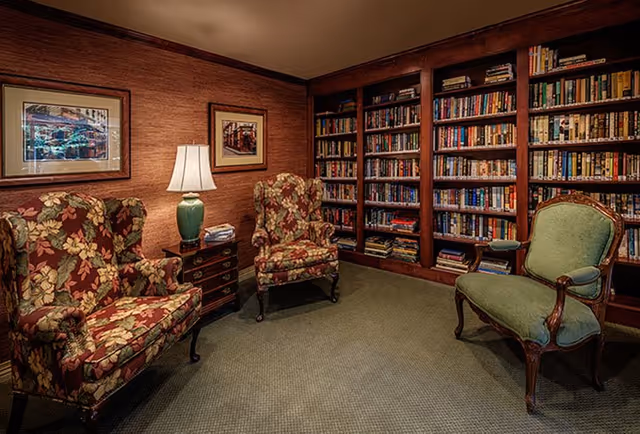 Cozy library room with two floral upholstered armchairs, one green upholstered armchair, a wooden side table with a green lamp, and a large wooden bookshelf filled with books. The walls are decorated with framed artwork.