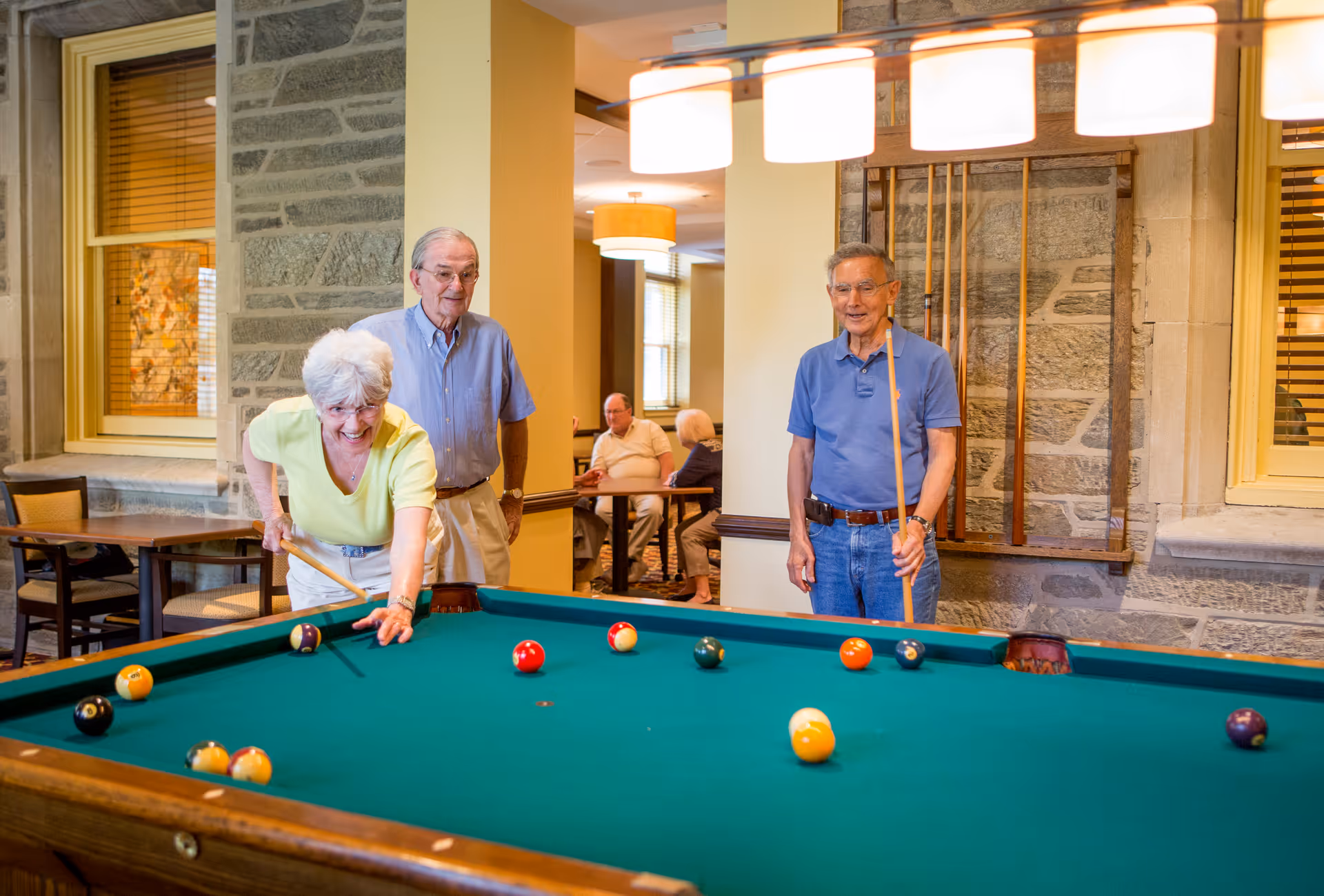 Three elderly people playing pool in a well-lit room with stone walls and wooden window frames. Two men stand near the pool table holding cues, while a woman is leaning over the table aiming to strike a ball. In the background, two other elderly people are seated at a table engaged in conversation.