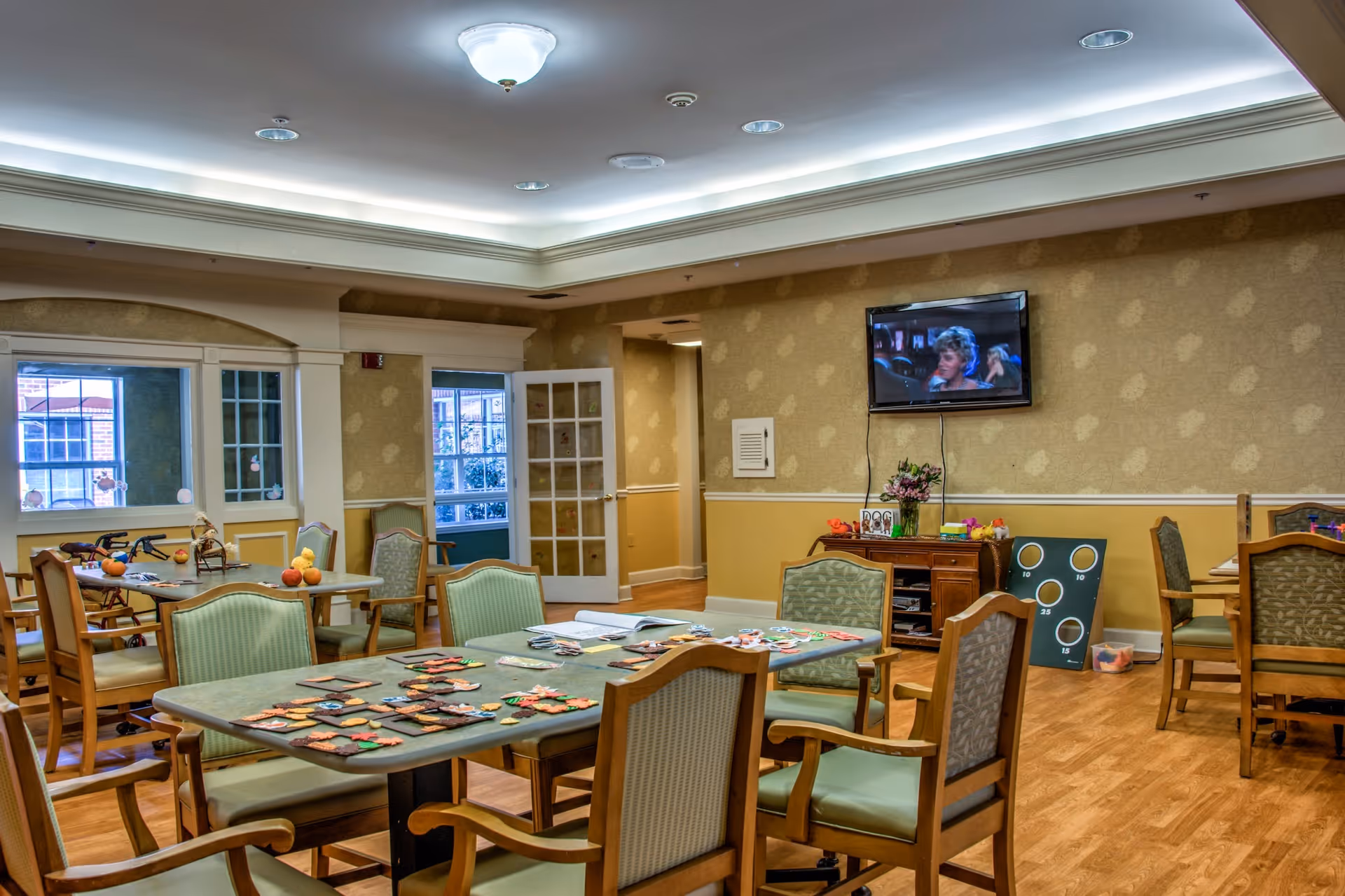 A well-lit common room with several tables and chairs arranged for group activities. On the tables are various craft materials and games. A flat-screen TV is mounted on the wall showing a scene from a movie. The room has beige walls with a patterned wallpaper and wooden flooring. There are windows and a glass door leading to another room.