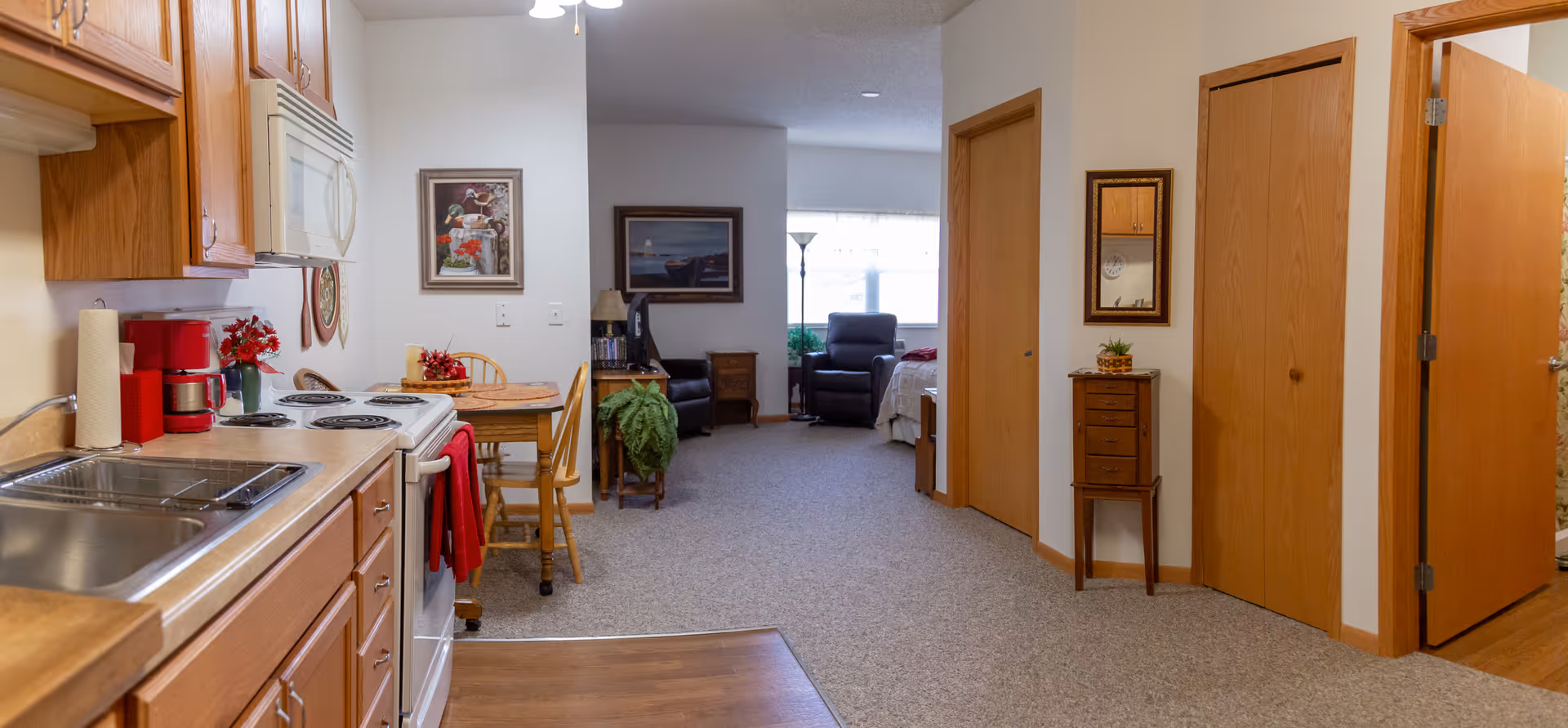 Interior view of a senior living apartment showing a kitchen area with wooden cabinets, a stove, microwave, and sink on the left. A small dining table with chairs is next to the kitchen. Beyond the dining area is a living space with armchairs, a floor lamp, and framed pictures on the walls. There are two wooden doors on the right side of the hallway, one partially open, and a small wooden cabinet with a mirror above it.