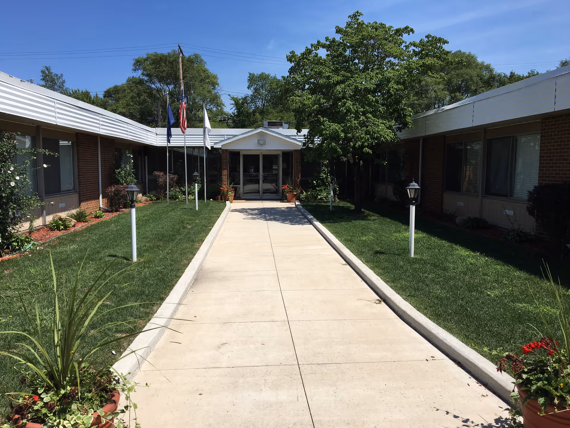 Concrete walkway leading to the entrance of a single-story brick senior living facility with flags and landscaped lawns.
