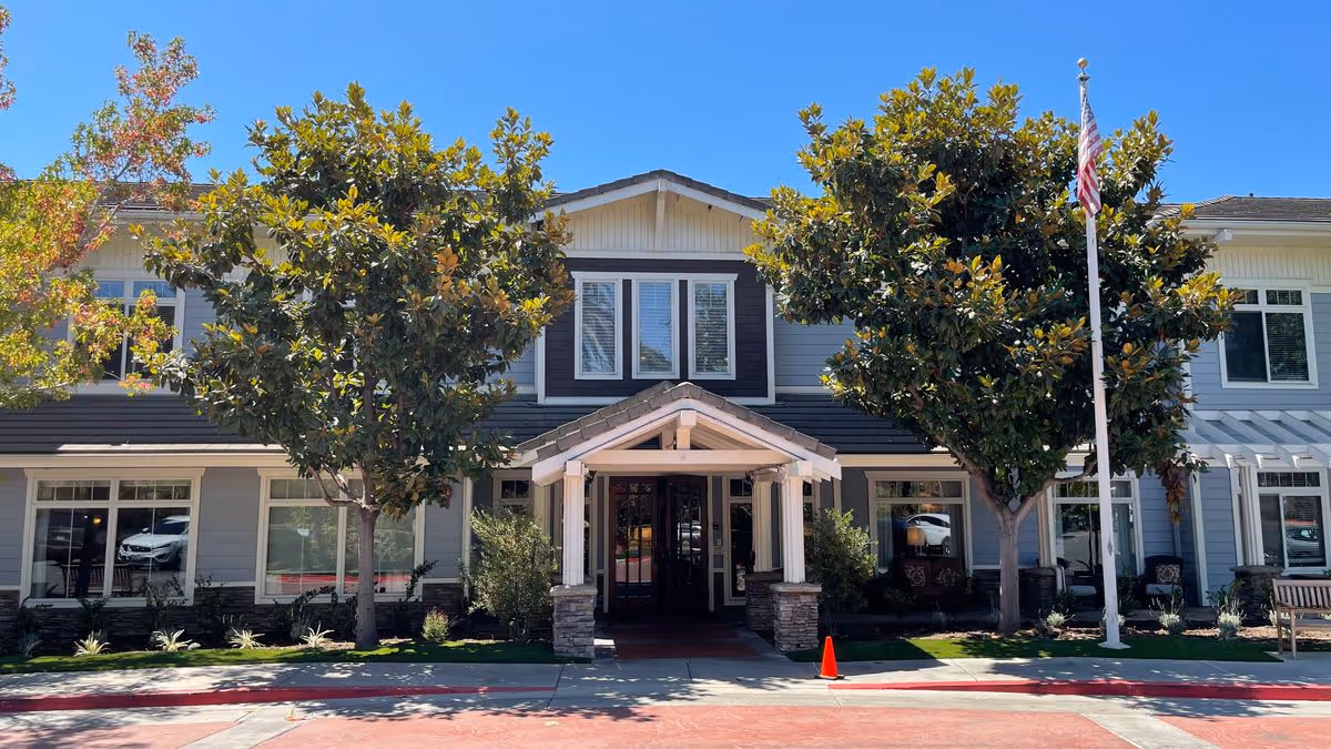 Front entrance of a two-story residential building with a covered portico, trees, and an American flag.