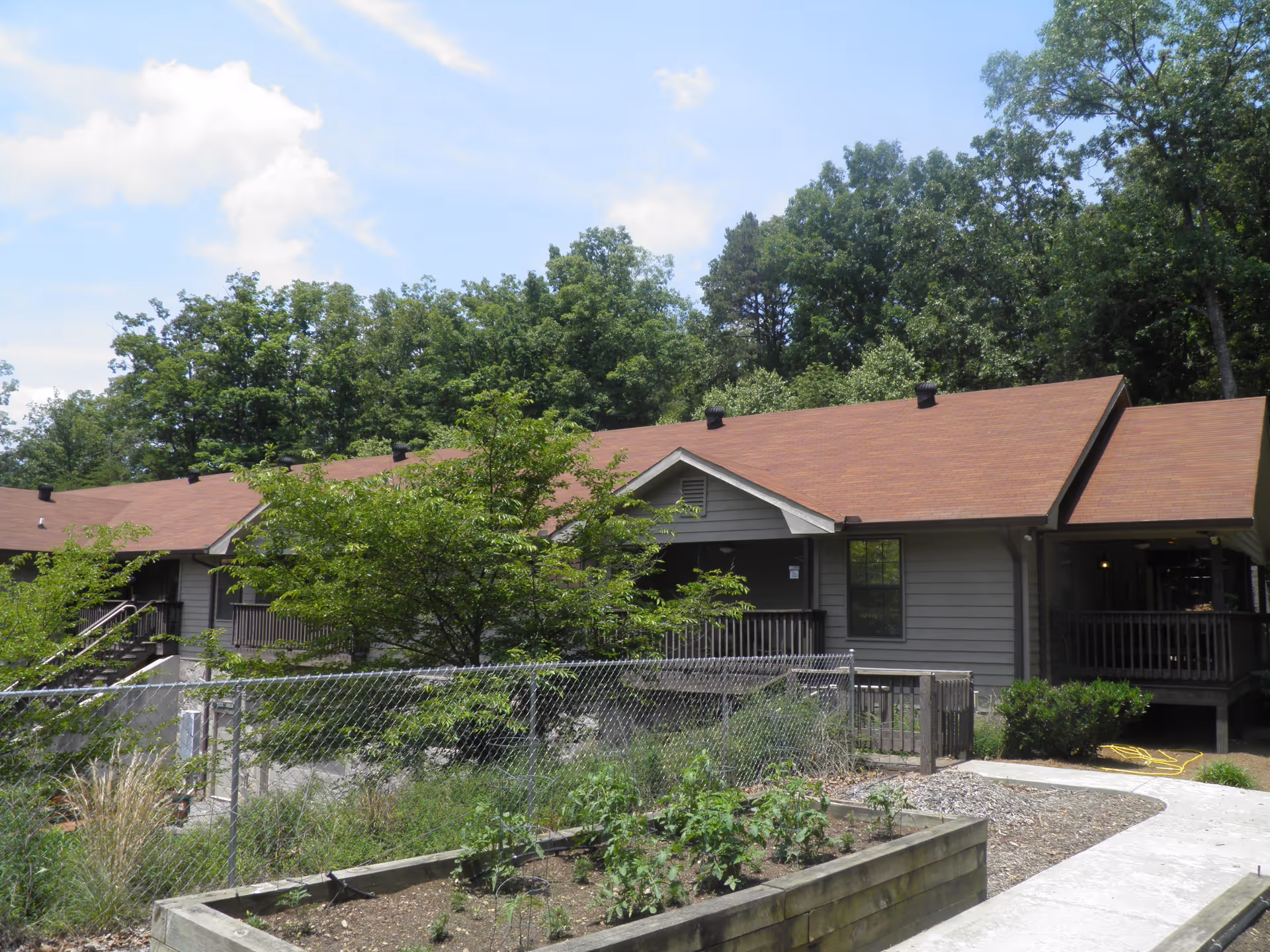Exterior view of a single-story building with a brown roof surrounded by trees and greenery. There is a chain-link fence enclosing a small garden area with plants, and a concrete pathway leading to the building's entrance.