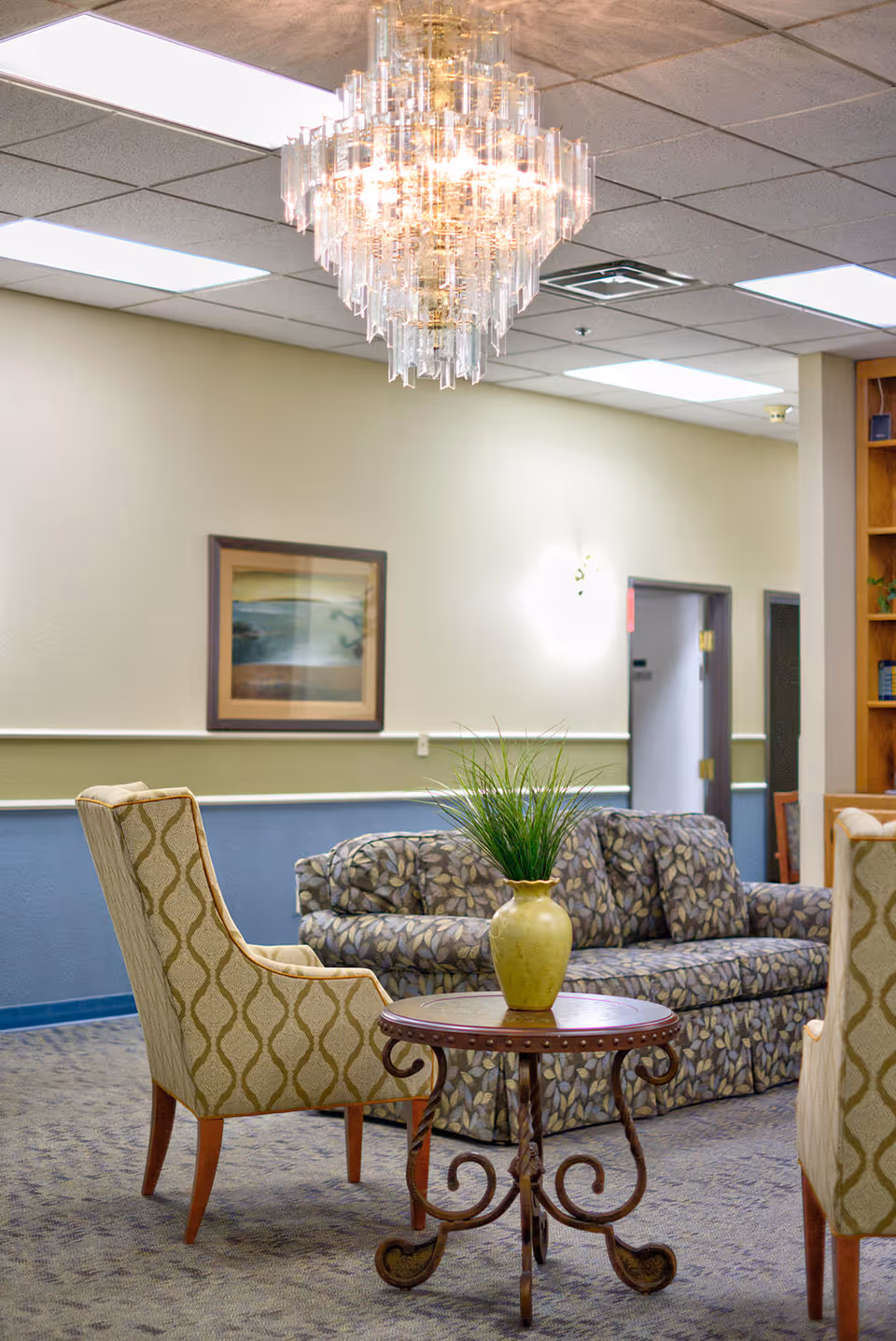 A seating lounge with patterned armchairs and a floral sofa around a small round table topped by a green vase, under a crystal chandelier.