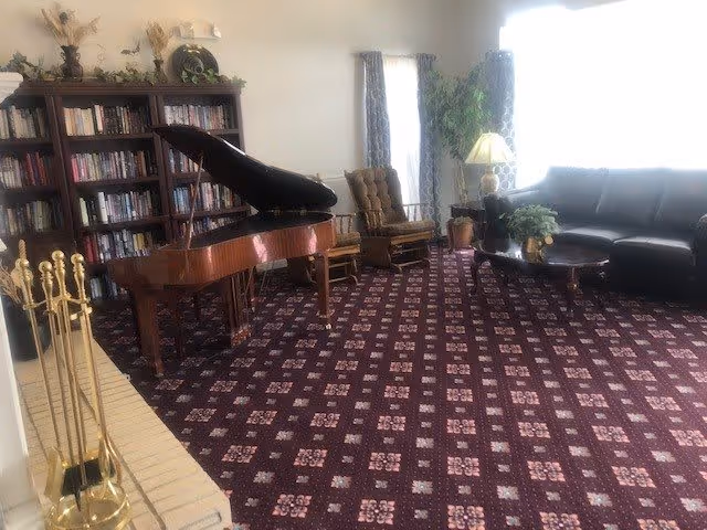 A cozy living room with a patterned carpet, a grand piano, a bookshelf filled with books, a fireplace with brass fireplace tools, a brown armchair, a black leather sofa, a wooden coffee table with a plant, and a floor lamp near a window with curtains.