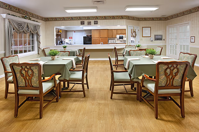 A dining room with several tables covered in green tablecloths, each surrounded by wooden chairs with cushioned seats and backs. Small potted plants serve as centerpieces on the tables. The room has wood flooring, beige walls with a floral border near the ceiling, and large windows with draped curtains. In the background, there is a kitchen area with wooden cabinets and appliances.