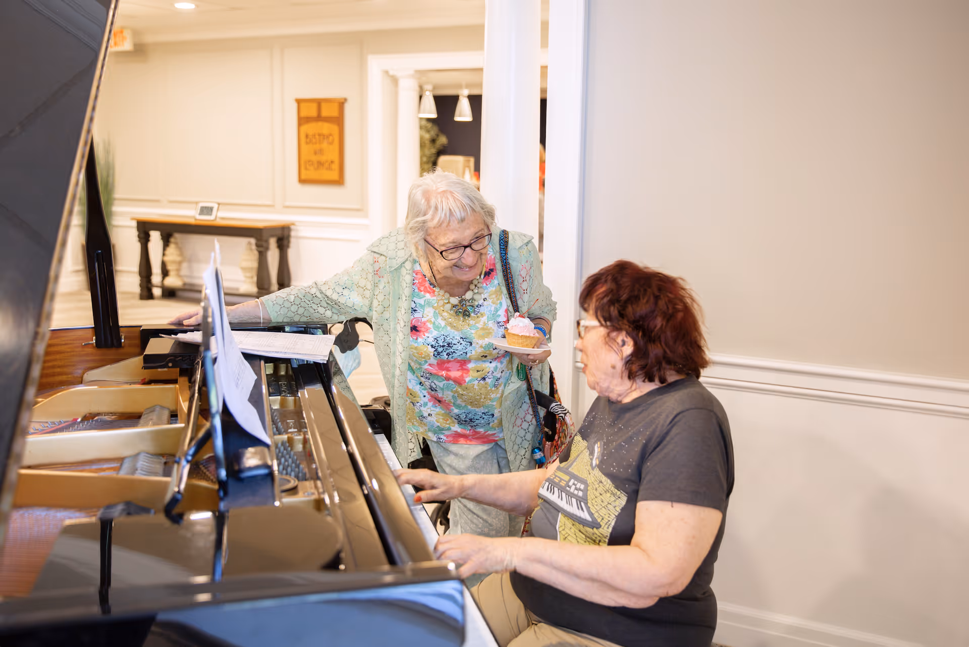 Two elderly women interacting by a grand piano in a well-lit room. One woman is seated playing the piano, while the other stands beside her holding a plate with a cupcake, smiling and engaging in conversation.