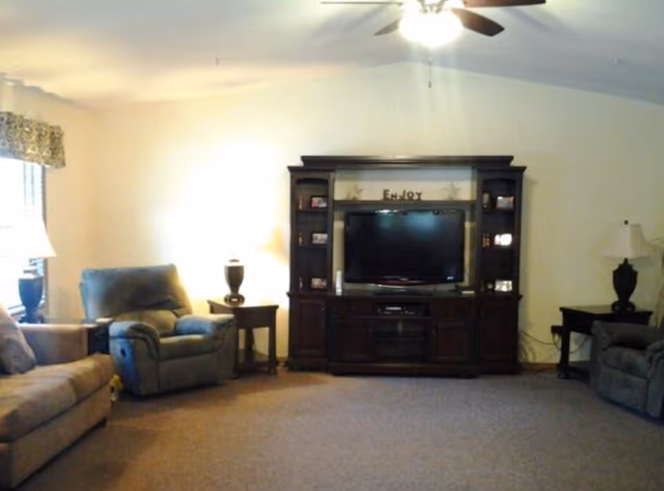 A living room with a beige sofa on the left, a blue recliner chair next to a wooden side table with a lamp, a large dark wood entertainment center with a flat-screen TV in the center, and another recliner chair on the right next to a small table with a lamp. The walls are light-colored and there is a ceiling fan with lights above.