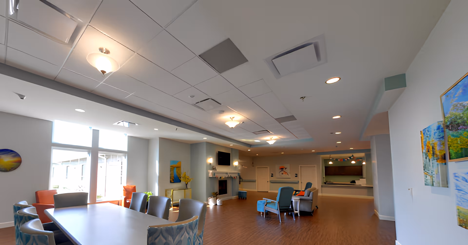 A spacious senior living common area with a large table and chairs in the foreground, comfortable seating including armchairs and a sofa near a fireplace with a TV mounted above it, bright windows letting in natural light, and colorful artwork on the walls.