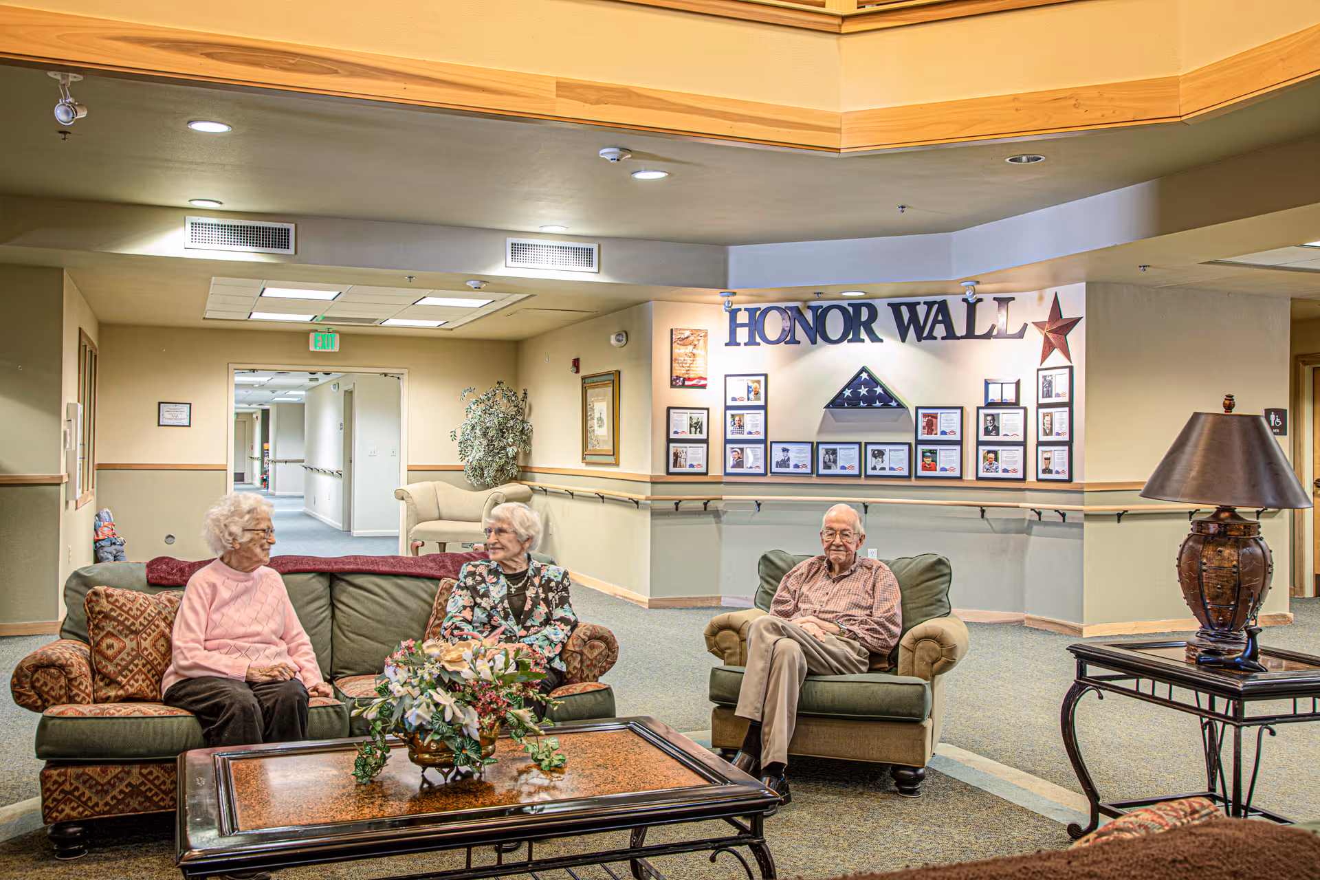 A cozy common area in a senior living facility with three elderly individuals seated on cushioned chairs and sofas. Behind them is a wall labeled 'HONOR WALL' displaying framed photos and a folded American flag. The room is warmly lit with a table lamp on a side table and a coffee table with a floral arrangement in the center.