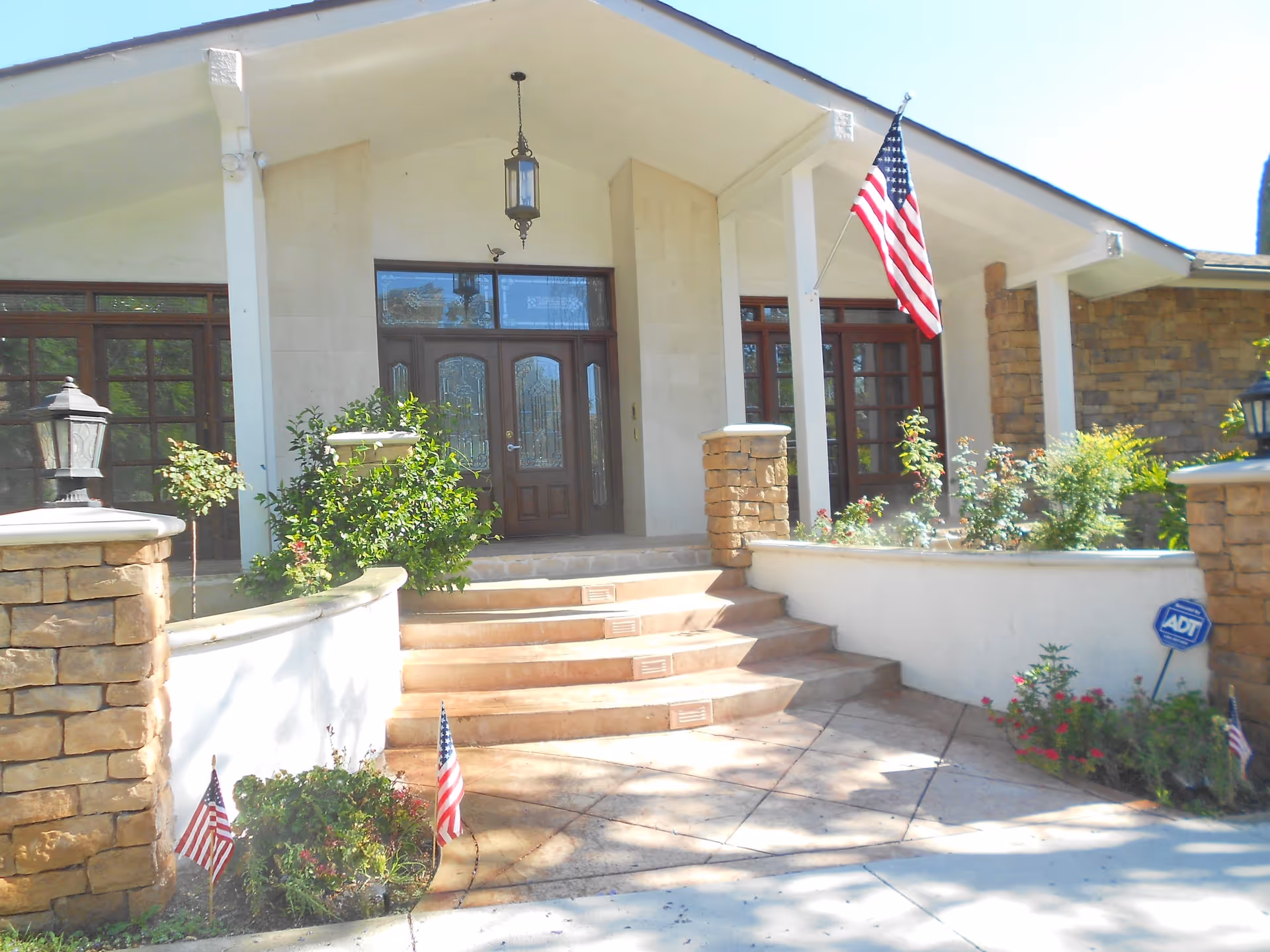 Front entrance of a building with stone pillars, steps leading up to double wooden doors with glass panels, an American flag mounted on the right side, and various plants and small American flags along the walkway.
