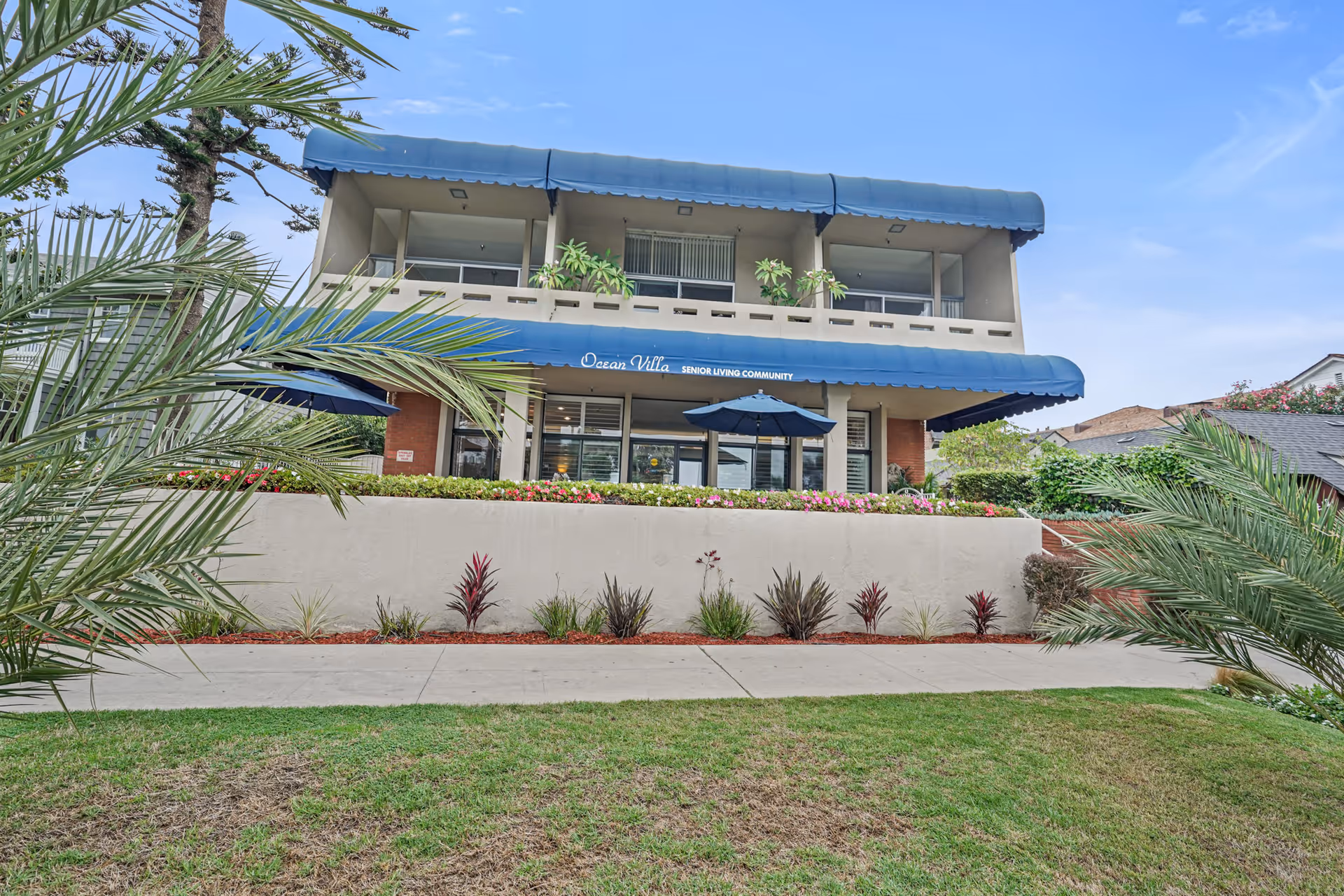 Exterior view of a two-story senior living community building named Ocean Villa with blue awnings, a balcony with plants, and a patio area with an umbrella. The building is surrounded by landscaped greenery and palm trees under a clear blue sky.