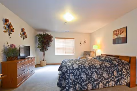 A furnished senior living bedroom with a large bed, dresser, TV, and window.