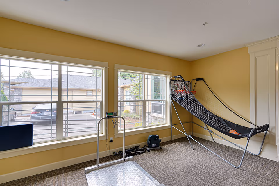 A bright room with large windows showing an outdoor parking area. Inside the room, there is an electronic scale platform and a basketball arcade game with a netted ramp and two basketballs. The walls are painted yellow and the floor is carpeted.