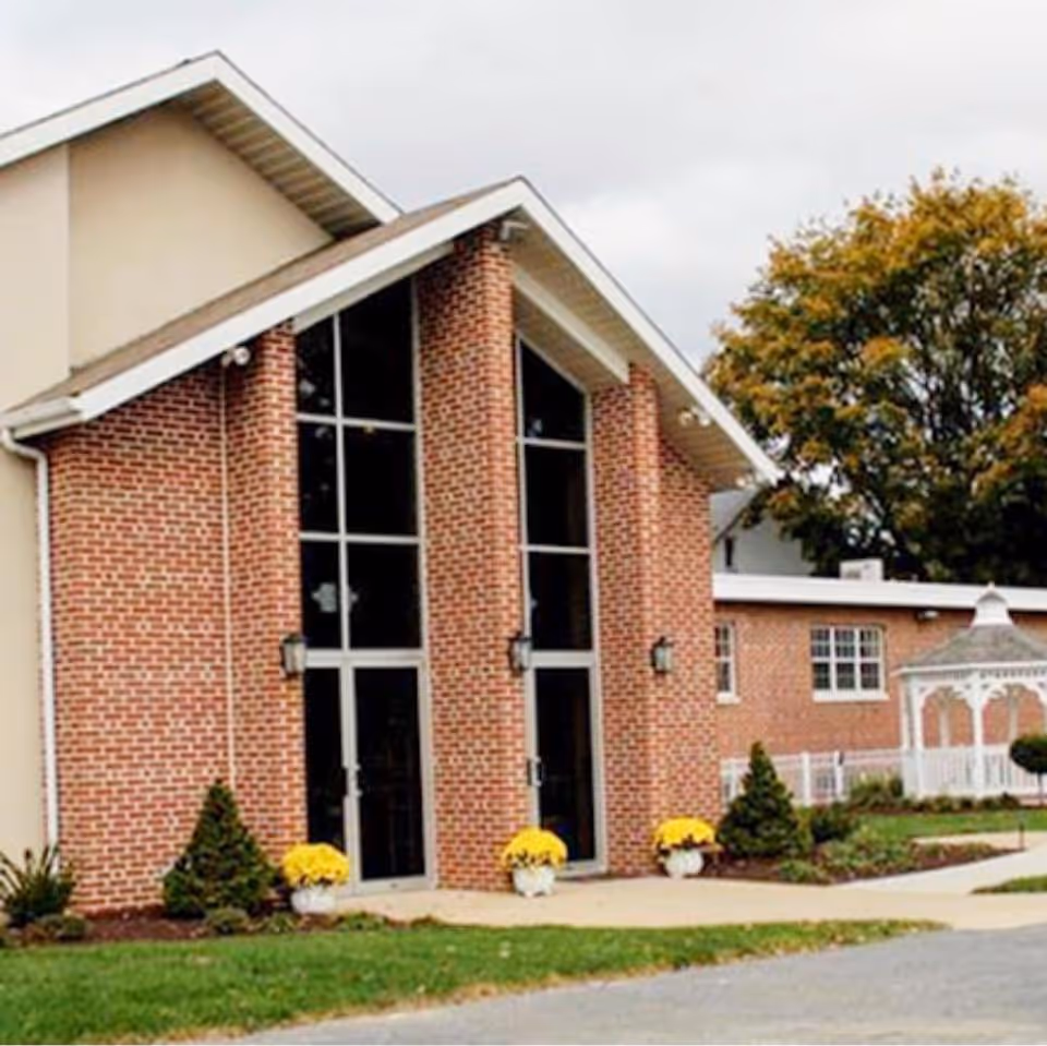 Exterior view of a brick building with large vertical windows and a peaked roof. There are small shrubs and yellow flowers in pots near the entrance, and a white gazebo is visible to the right. Trees with autumn foliage are in the background.